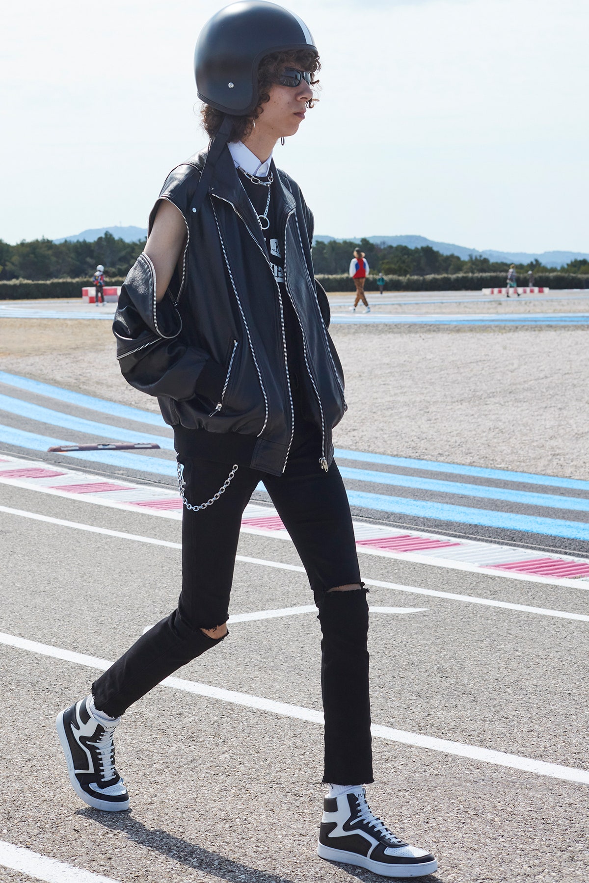 A model wearing black pants a black leather jacket black and white celine sneakers and a black motorcycle helmet