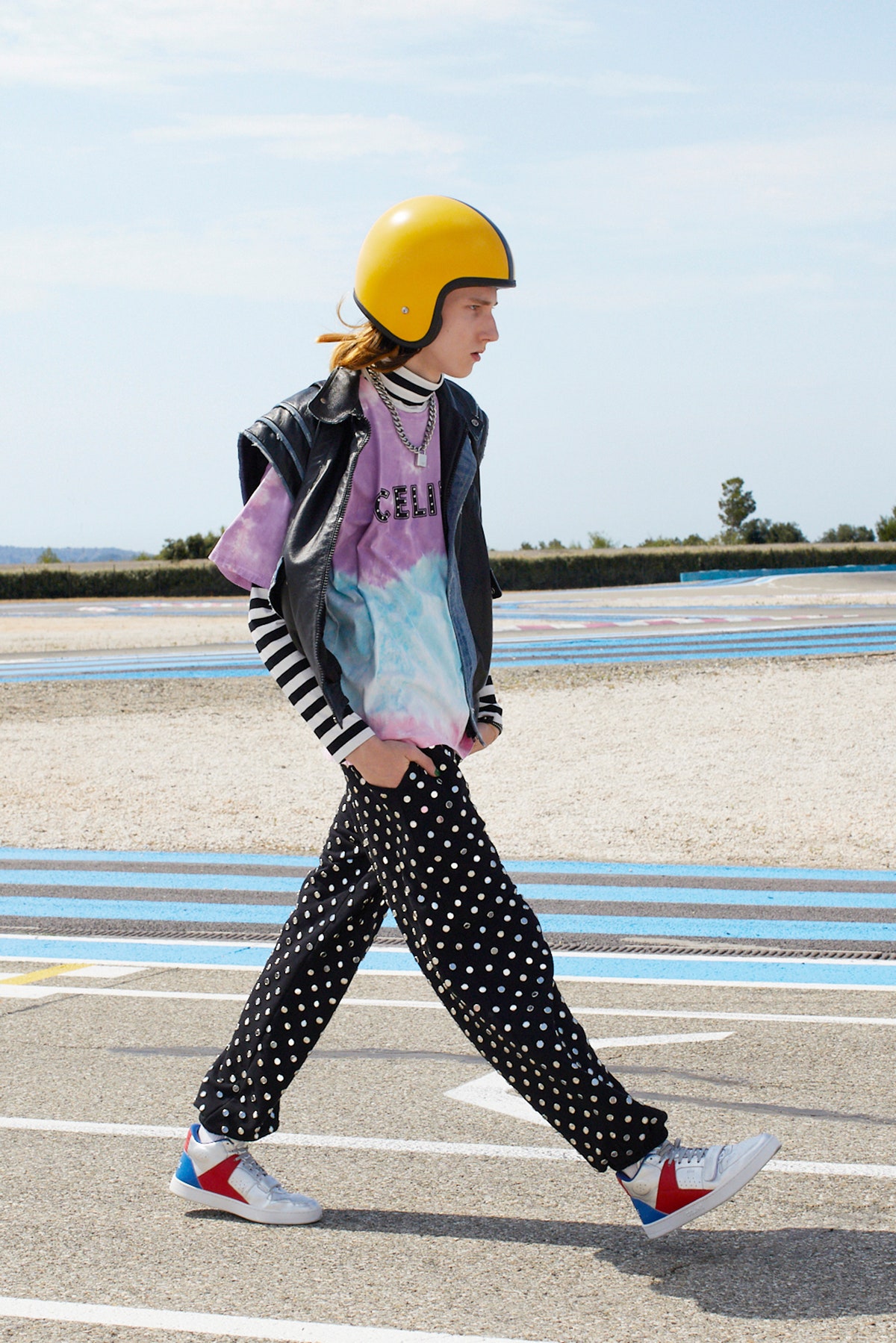 A model wearing polka dot pants a tie dye shirt red and blue celine sneakers and a yellow motorcycle helmet