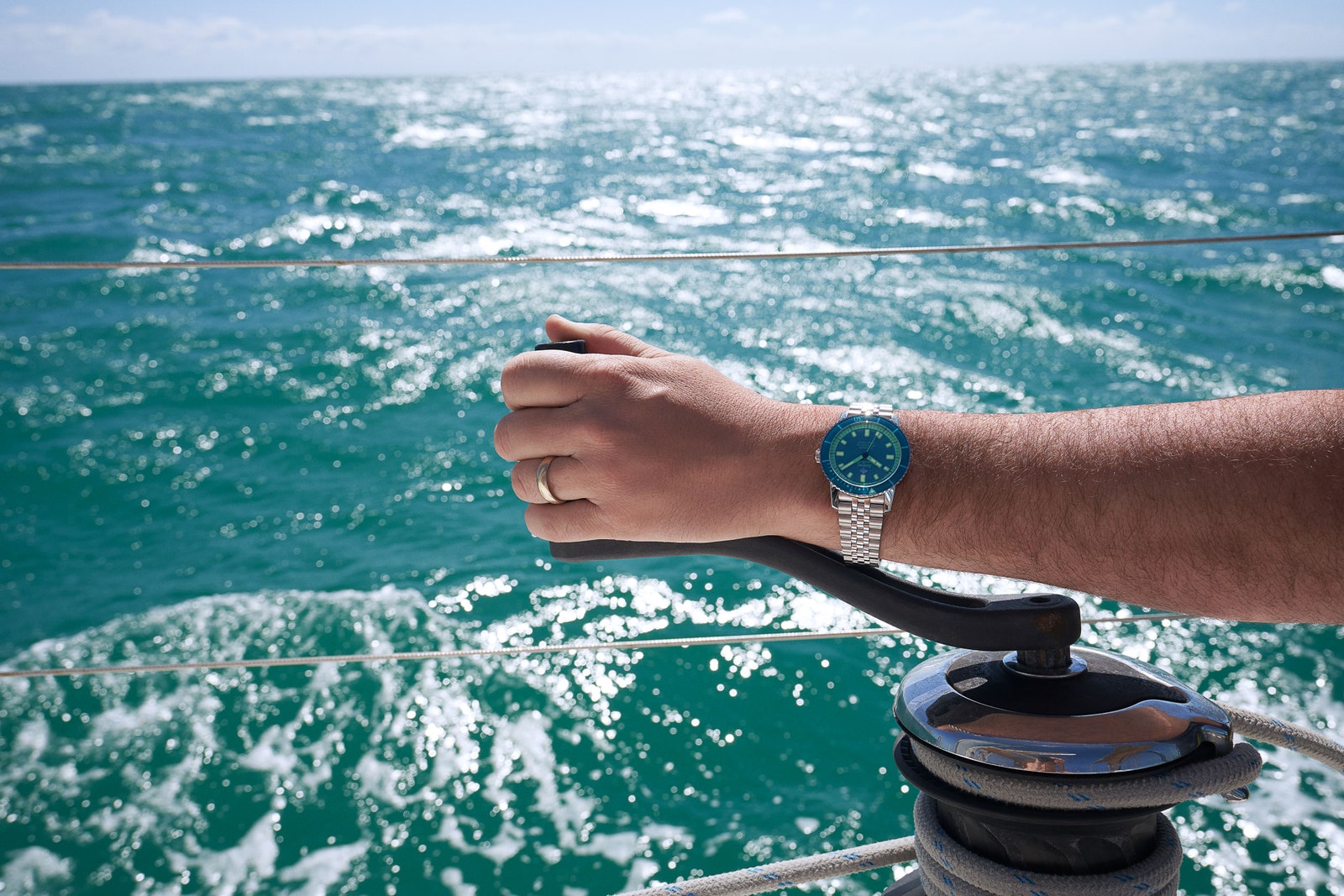 A hand wearing a steel watch with blue and green accents cranking a lever on a boat on the ocean