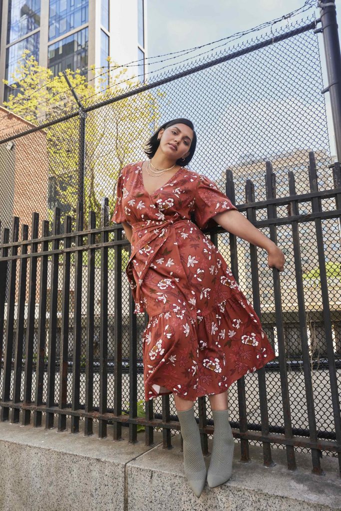 Woman standing against fence in red floral midi dress from ELOQUII elements.