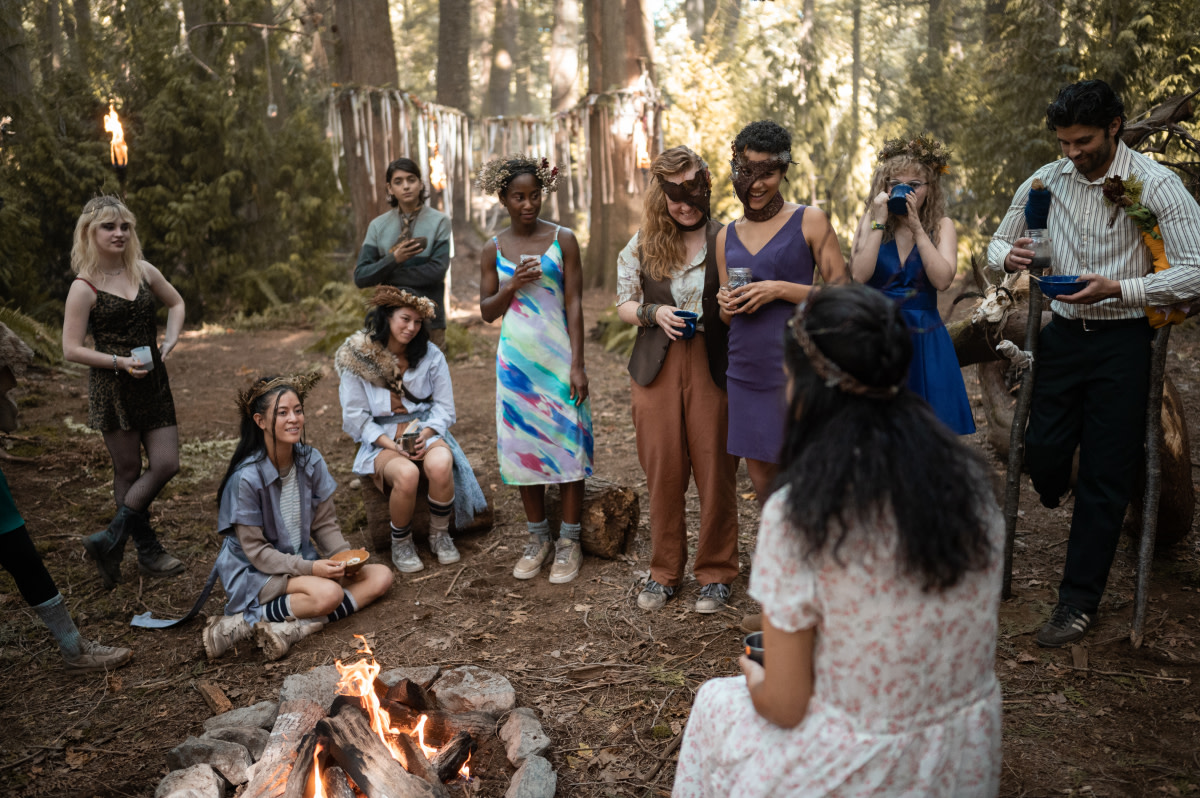 Doomcoming L–R: Natalie in leopard print, two Yellowjacket teammates, Javi (Luciano Leroux), Akilah, Van (Liv Hewson), Taissa (Jasmin Savoy Brown), Misty (Samantha Hanratty) and Coach Ben (Steven Krueger) look on as Lottie (foreground) sings Seal's 1994 hit 'Kiss From a Rose.'